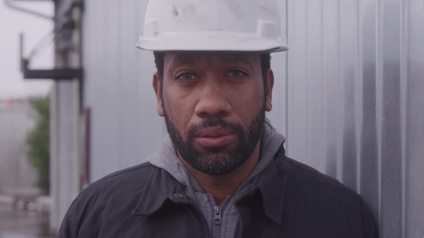 A man wearing a hard hat stands in front of a building and turns his head slowly