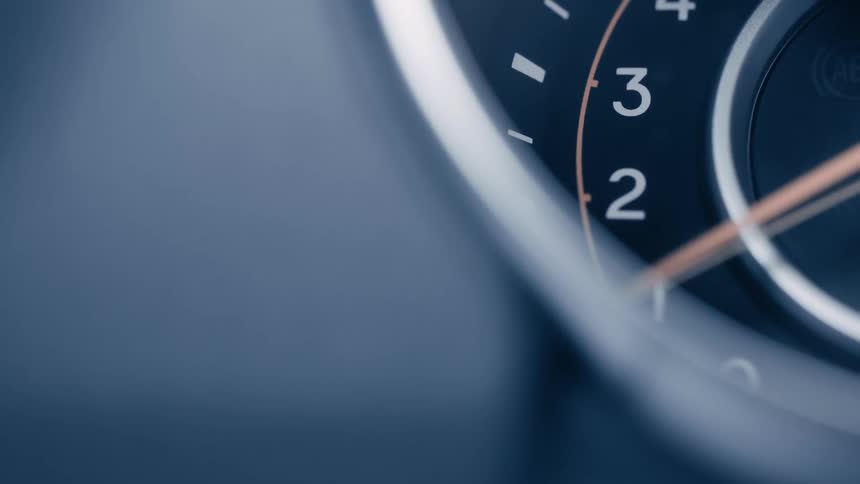 A closeup angled view of a cars speedometer or tachometer with the orange needle moving up and down with part of the dashboard visible on the left all in shades of dark blue and grey