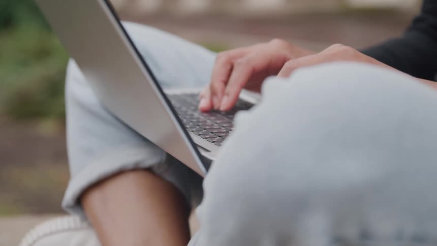 Close up of a person sitting outdoors typing on thier laptop