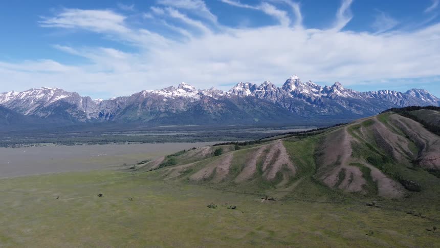 Snowcapped mountain range stretches beneath a blue sky with wispy clouds and surrounding green plains