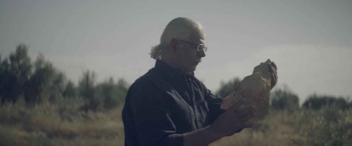 An elderly man picks up a large rock outdoors