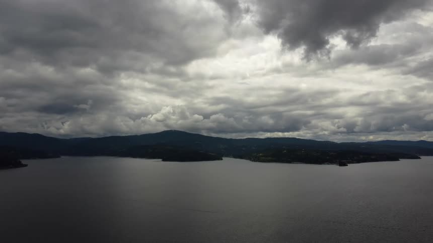 The video shows an expansive aerial view of a dark lake or body of water surrounded by mountainous terrain under a heavily overcast sky the clouds are dark and dramatic suggesting an impending storm or just a very cloudy day