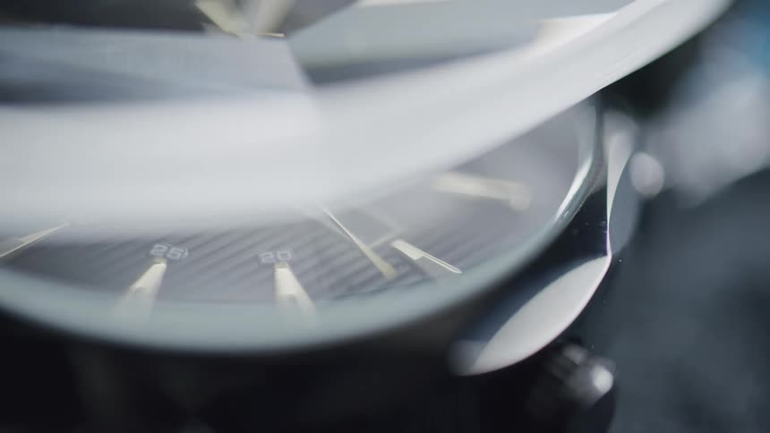 A close view shows a wristwatch with a dark face and light colored minute and second markers with light reflecting off the glass and watch casing