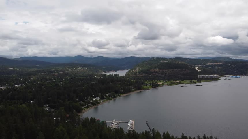 The video offers an expansive aerial view of a large lake surrounded by lush green forests and mountains under a cloudy sky boats are docked along the shoreline adding a touch of human presence to the natural landscape