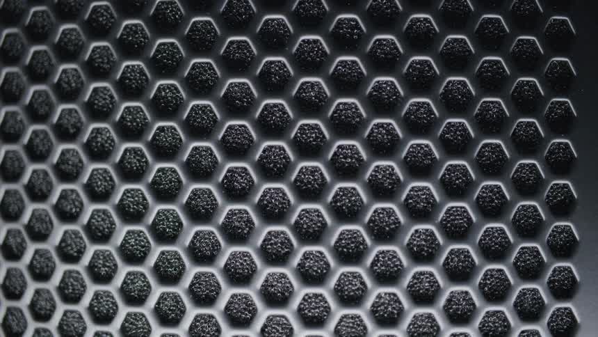 A close view of a textured surface shows a repeating pattern of dark granular hexagons set within a lighter grey honeycomb grid under shifting lights