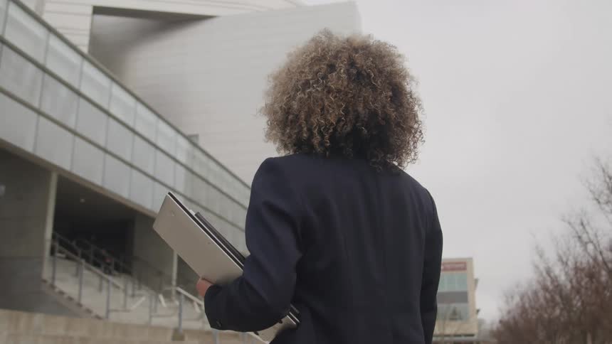 A young adult woman with curly hair wearing a dark blazer stands holding a laptop and looking towards a large modern building with stairs