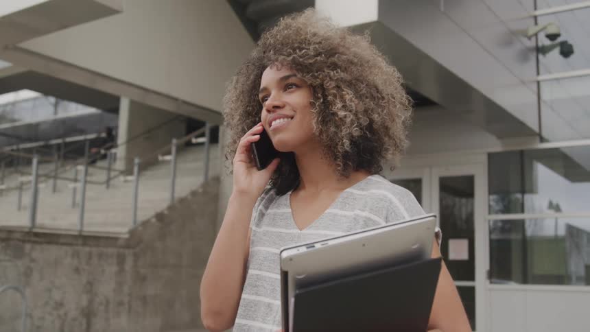 Smiling woman talks on phone holding laptop and notebook outside