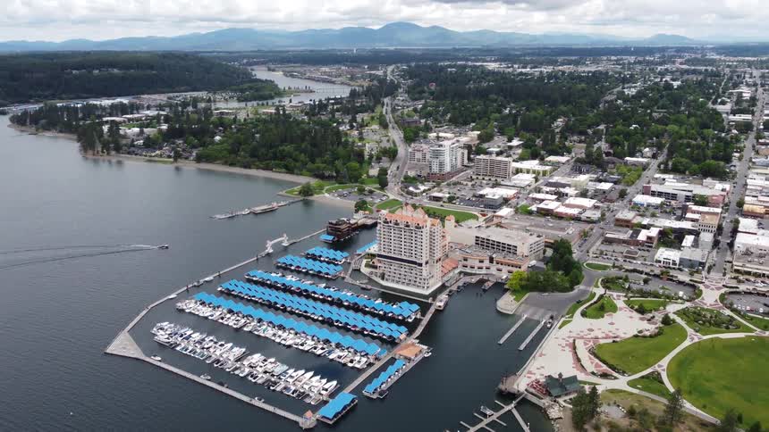 The video presents a highangle aerial view of a lakeside town featuring a prominent marina filled with numerous boats docked under blue covers a large building with a distinctive orange roof stands near the water surrounded by other town buildings