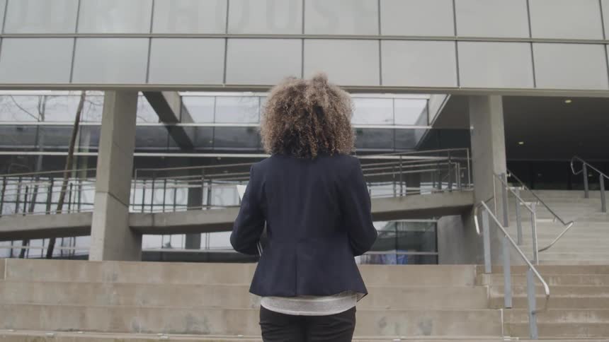 A woman with large curly hair viewed from behind stands at the bottom of a wide concrete staircase looking up at a modern glass building