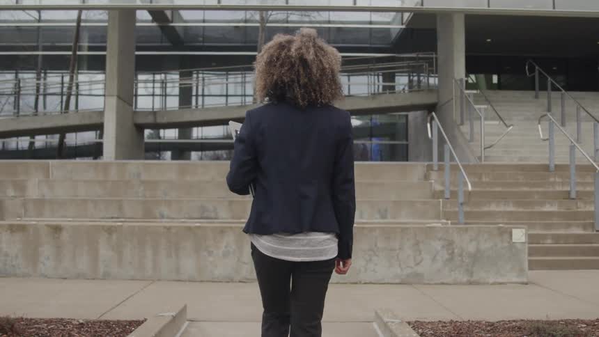 A woman with curly brown hair walks away from the camera towards a large modern building with glass windows and concrete stairs