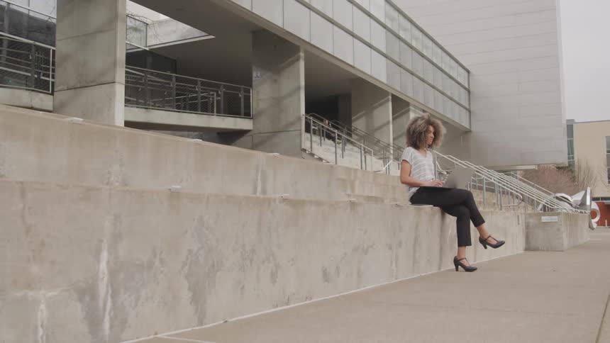 A young woman sitting on a concrete slab in front of a building working on her laptop