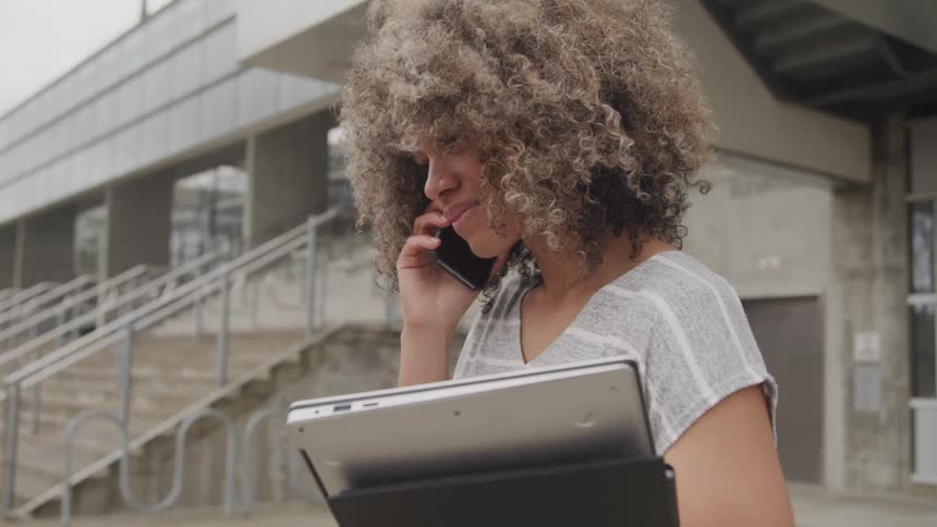 Woman talking on the phone while holding a laptop outdoors