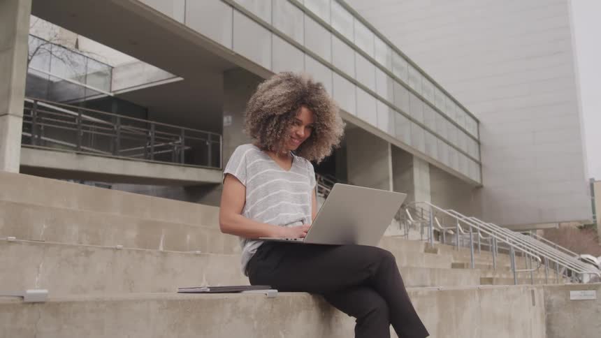 A young black woman uses a laptop while sitting on a pavement outdoors during daytime