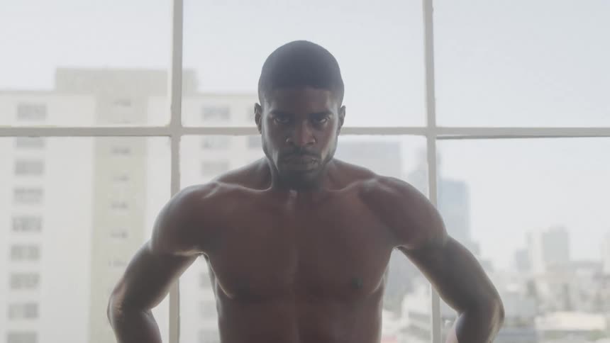 The video shows a man from the chest up shirtless with a muscular build he is looking directly at the camera a large window is behind him through which buildings can be seen in the background the lighting is bright possibly from the window and cas