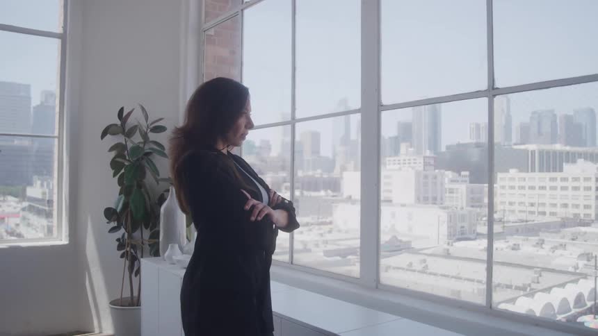 Professional woman with arms crossed looking out large office window to city below