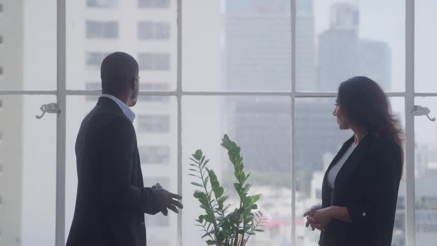 Two coworkers are talking in the office by the window