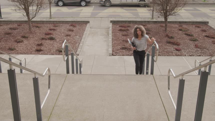A woman with light colored curly hair wearing a striped shirt and dark pants walks up a wide concrete staircase with metal handrails carrying a laptop with a paved area small trees and parked vehicles in the background