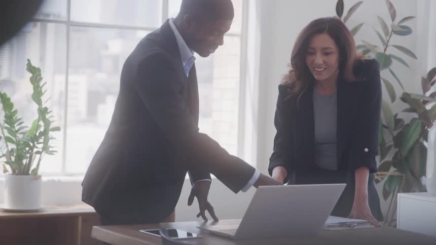 The video shows two professionals a man and a woman standing around a desk with a laptop on it the man on the left is wearing a suit and appears to be pointing at something on the laptop or desk while the woman also in a suit is looking at laptop