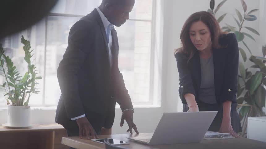 Two people in an office looking at a laptop presentation with the woman explaining to the man both are wearing suits