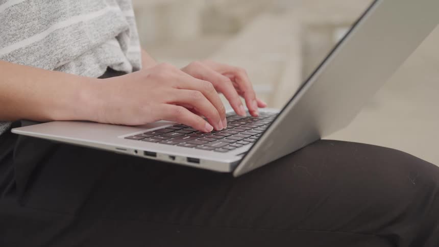 A person is seen from the waist up typing on a silver laptop that rests on their lap while wearing a grey striped shirt and dark pants