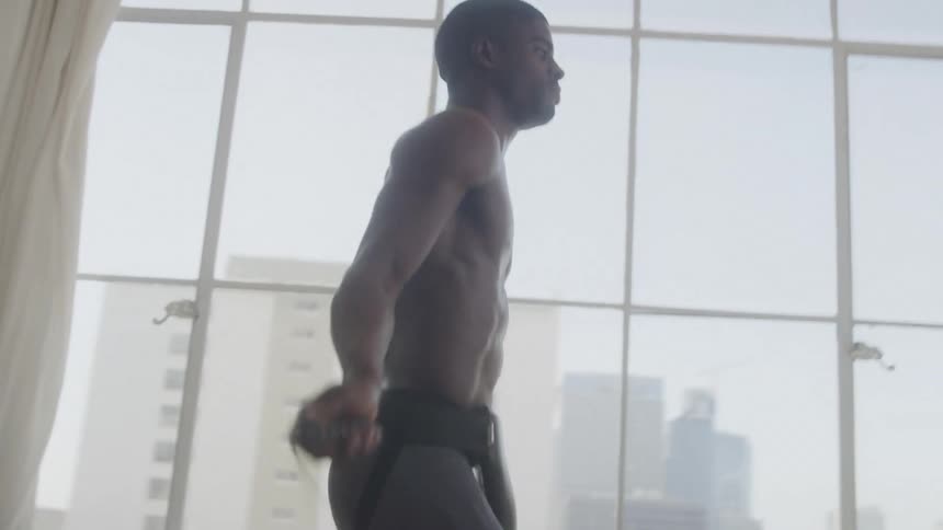 The video shows a shirtless man exercising in a bright room with large windows overlooking a city he is jumping rope with a serious and focused expression