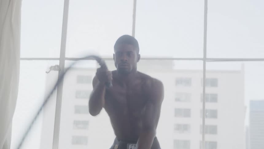 A fit darkskinned man in dark athletic wear intensely exercising with battle ropes in a welllit indoor space with large windows in the background