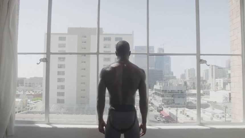 A shirtless man standing in front of glass windows looking out over the city from above