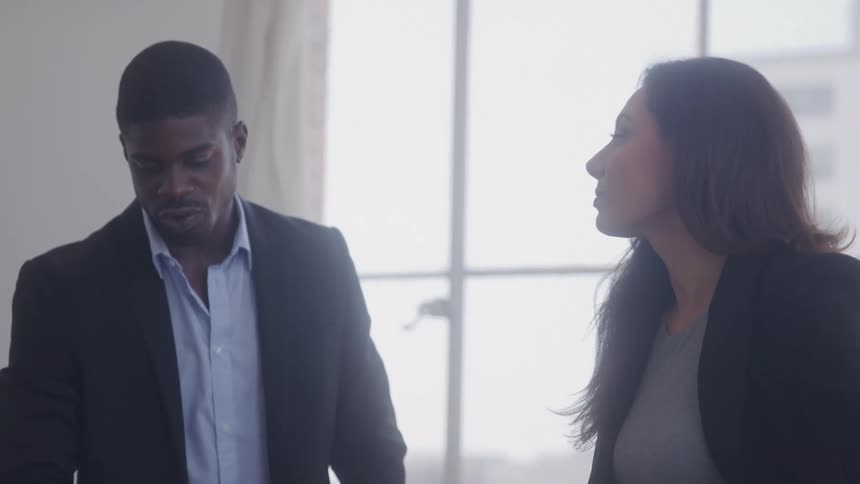 A male and female coworker converse while standing in an office during daytime