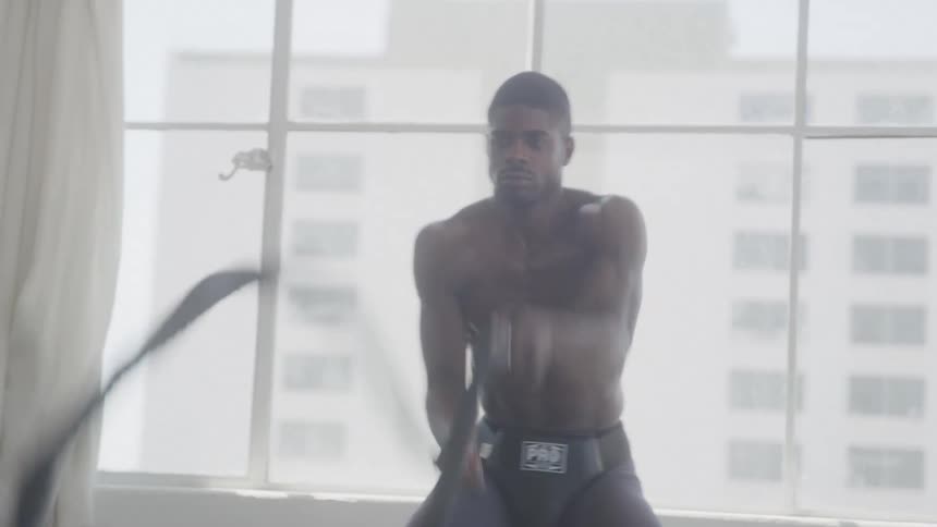 A shirtless man intensely exercises with battle ropes in a sunlit room