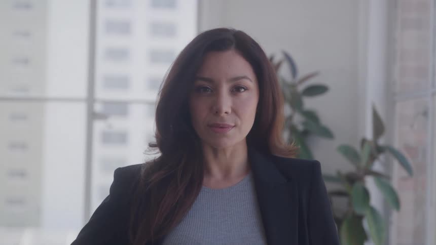 A confident businesswoman stands in a modern office with a neutral expression
