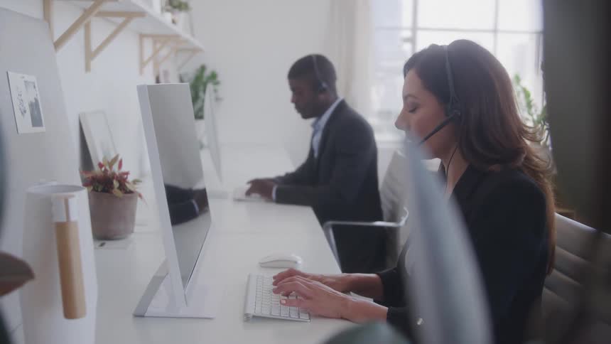 The video shows a busy office environment with two people working at computers in the foreground a woman wearing a headset is typing on a keyboard while in the background a man is also working at a computer both appear to be engaged in customer servi