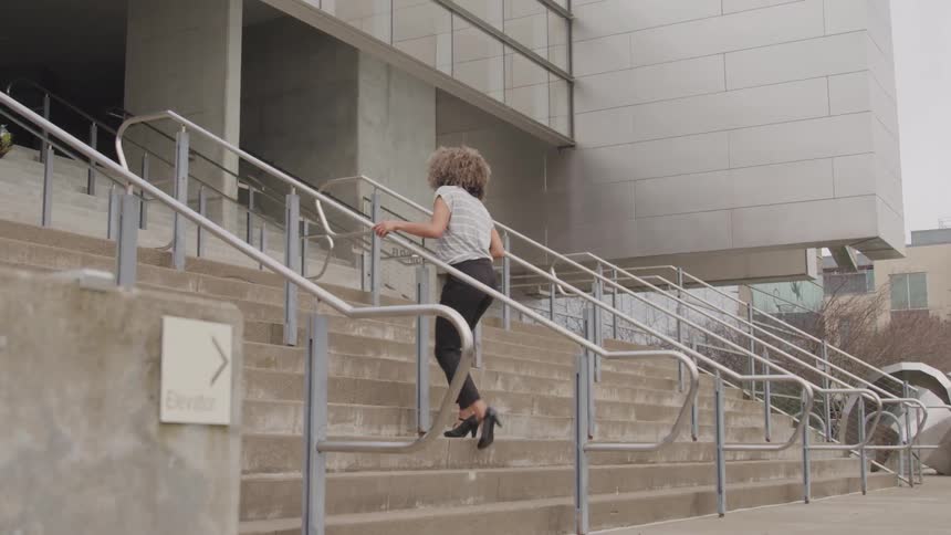 The video shows a woman with curly hair walking up a large outdoor concrete staircase she is wearing a white striped top dark pants and black heels and is using the handrail to ascend the stairs