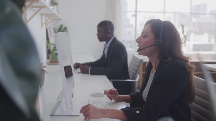 A man and a woman both colleagues sit at their desks facing computers wearing suits and headphones talking to customers inside the office at midday