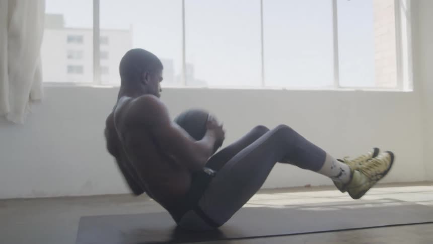 A male athlete performs a seated trunk rotation exercise with a ball while sitting on a mat indoors