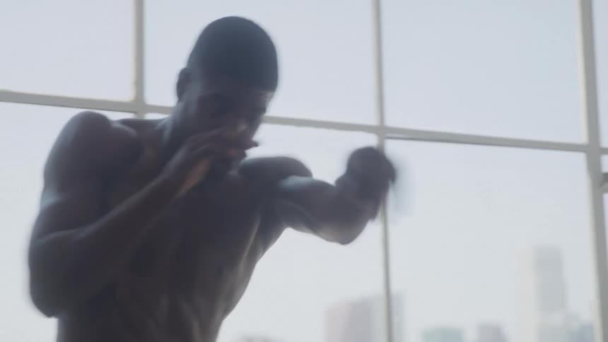 A shirtless man shadowboxes indoors at midday