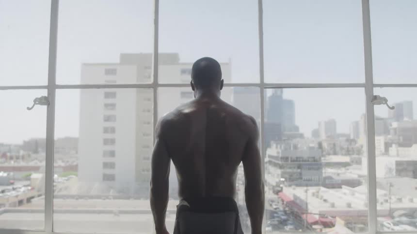 A shirtless young man standing and looking out a glass window revealing the cityscape view at midday