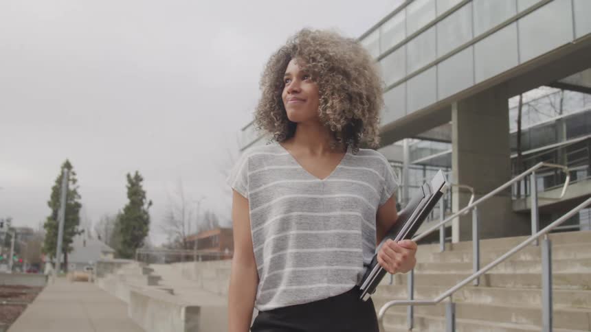 Woman with curly hair walking on the sidewalk next to a building and carrying folders of documents