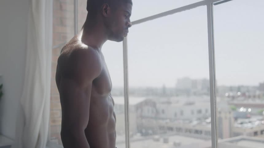The video shows a shirtless muscular man standing in profile looking out of a large window through the window a cityscape can be seen in the background under bright daylight the man appears to be contemplative or observing something outside