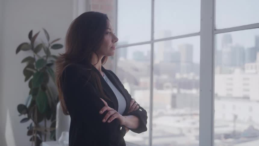 A thoughtful businesswoman wearing a black blazer and a white shirt is standing by a large window with her arms crossed looking out at a blurry cityscape