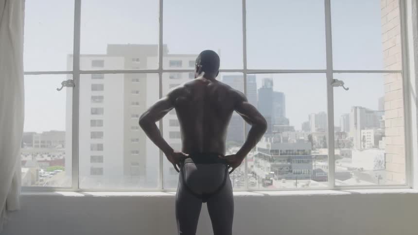 A muscular man standing in front of a window looking at the city view
