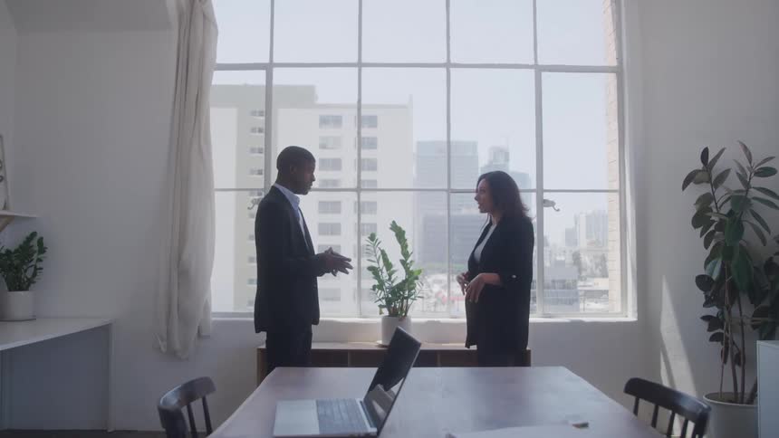 Two elegantly dressed business professionals are engaged in a conversation while standing in front of large windows overlooking a city skyline within a modern office space