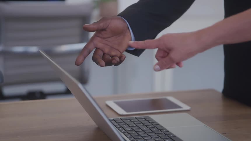 A closeup shot showing two hands one lightskinned and one darkskinned pointing towards a laptop screen on a wooden desk