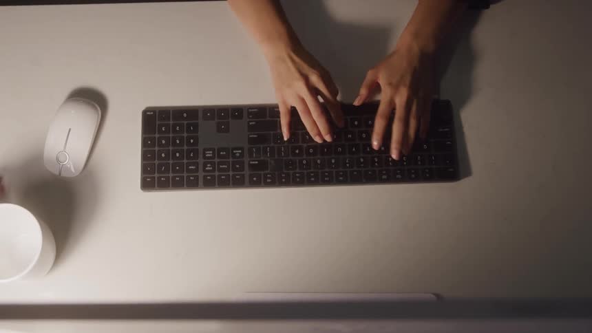 The video shows an overhead shot of a persons hands typing on a black keyboard positioned on a white desk a white computer mouse and a white mug are also visible to the left of the keyboard
