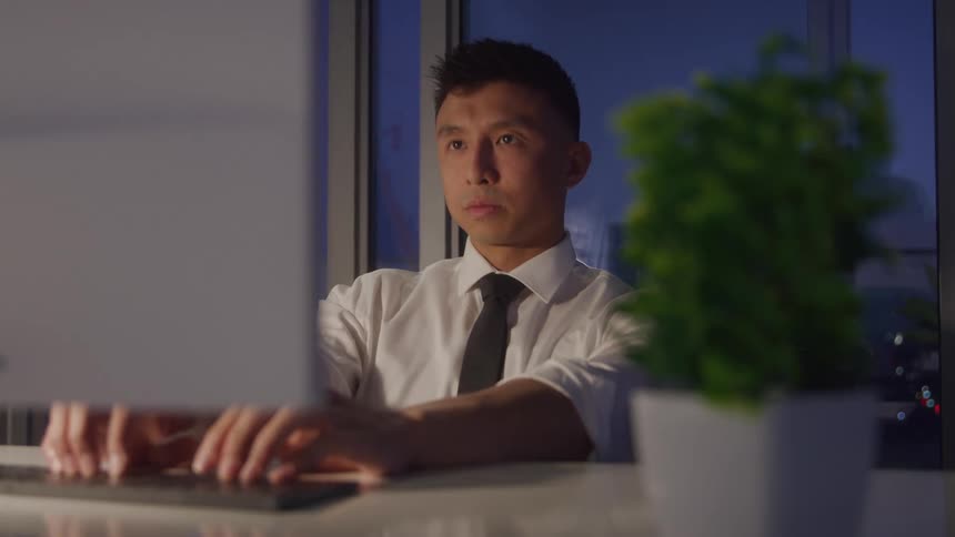 A young male worker uses a computer at a table in an office at night