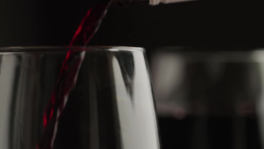 In a dramatic closeup shot a steady stream of rich red wine is poured from an offscreen bottle flowing elegantly into a clear wine glass against a dark moody background