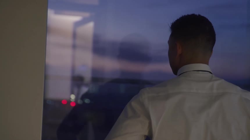 A man in a white collared shirt and dark tie stands with his back to the viewer looking out a large window at a dim blurry evening scene with distant lights