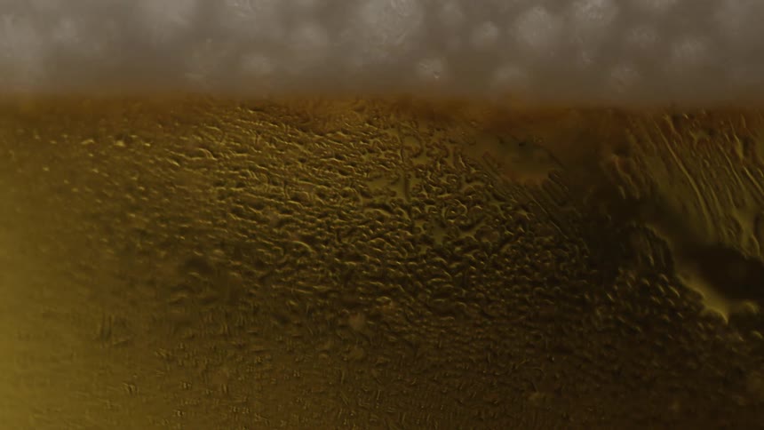 An extreme closeup focuses on the textured condensation covering a glass of golden beer with a wave of liquid visible on the inside and a soft white foam head at the top as bubbles rise to the top