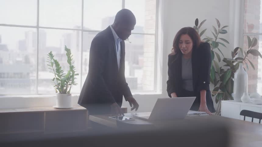 A man in a dark suit stands next to a woman in a dark blazer both looking at a laptop on a wooden table with large windows and plants in the background