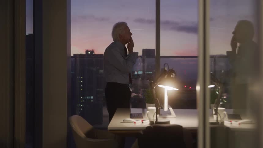An elderly man with white hair wearing a shirt and dark pants is standing by a large window overlooking a cityscape at dusk he appears to be talking on the phone holding it to his ear