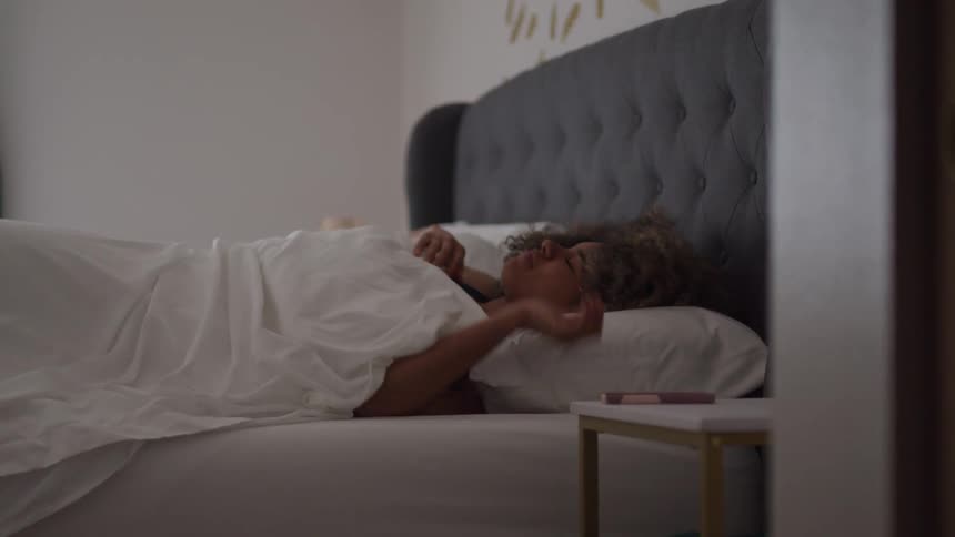 A woman lies in bed under white sheets staring thoughtfully toward a nightstand with a phone